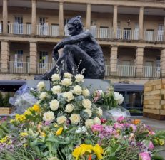 Statue of a woman sat down, resting one arm on her knee. Colourful flowers laid out at the bottom of the statue.