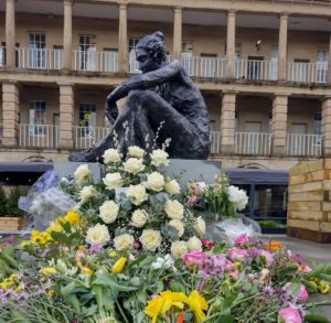 Statue of a woman sat down, resting one arm on her knee. Colourful flowers laid out at the bottom of the statue.