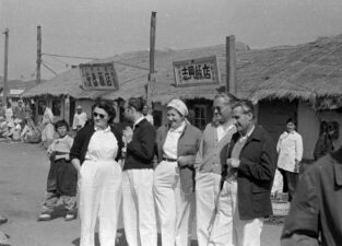 Black and white photograph. In the centre of the image stands five adults, two women and three men. Behind them are houses with thatched roofs.