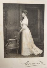 Black and white photograph with a woman standing in the centre. She is wearing a long white dress and is standing to the left in profile. She is holding onto a chair which is highly decorative.