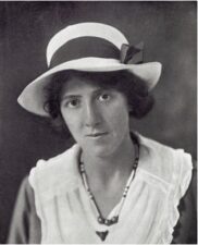 Black and white photograph of a young woman looking directly at the camera. She has a serious express and wears a white hat.
