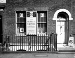 Black and white photograph of the bottom of a Victorian looking house. On the right is the front door and to the left large windows. There are black railings at the front of the house. On the window there is a sign which reads 'The Mother's Clinic'.