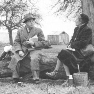 Black and white photograph of a man and woman sat on a log in a field. They are turned towards each other as if in conversation.